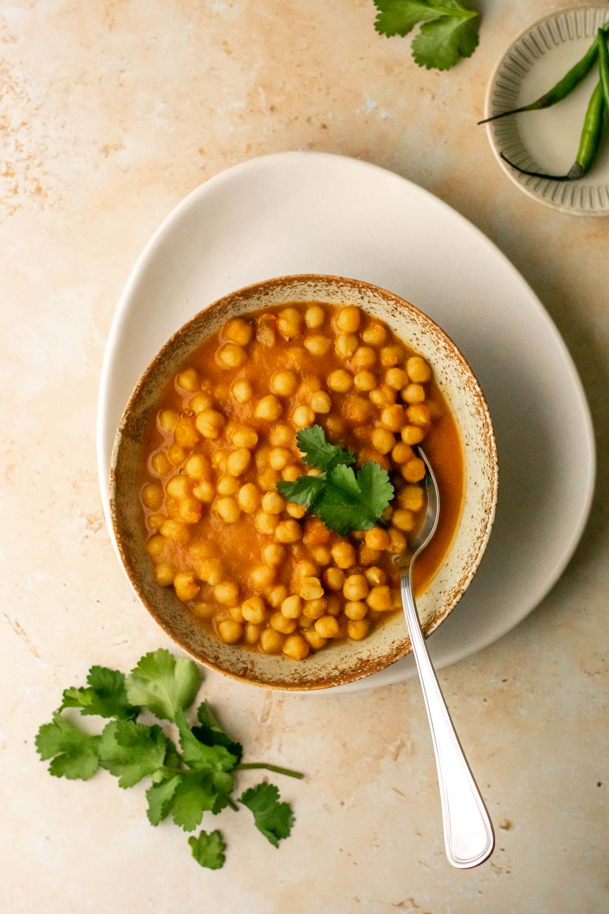 a bowl of chickpea curry on a plate served with coriander and fresh green chillies