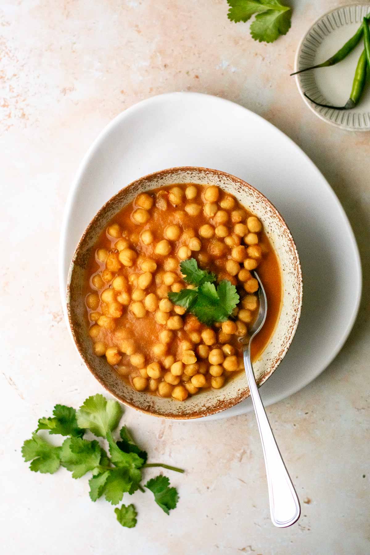 a bowl of chickpea curry on a plate served with coriander and fresh green chillies