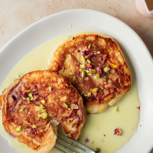two rasmalai pancakes on a plate with a fork, served in sweet saffron milk